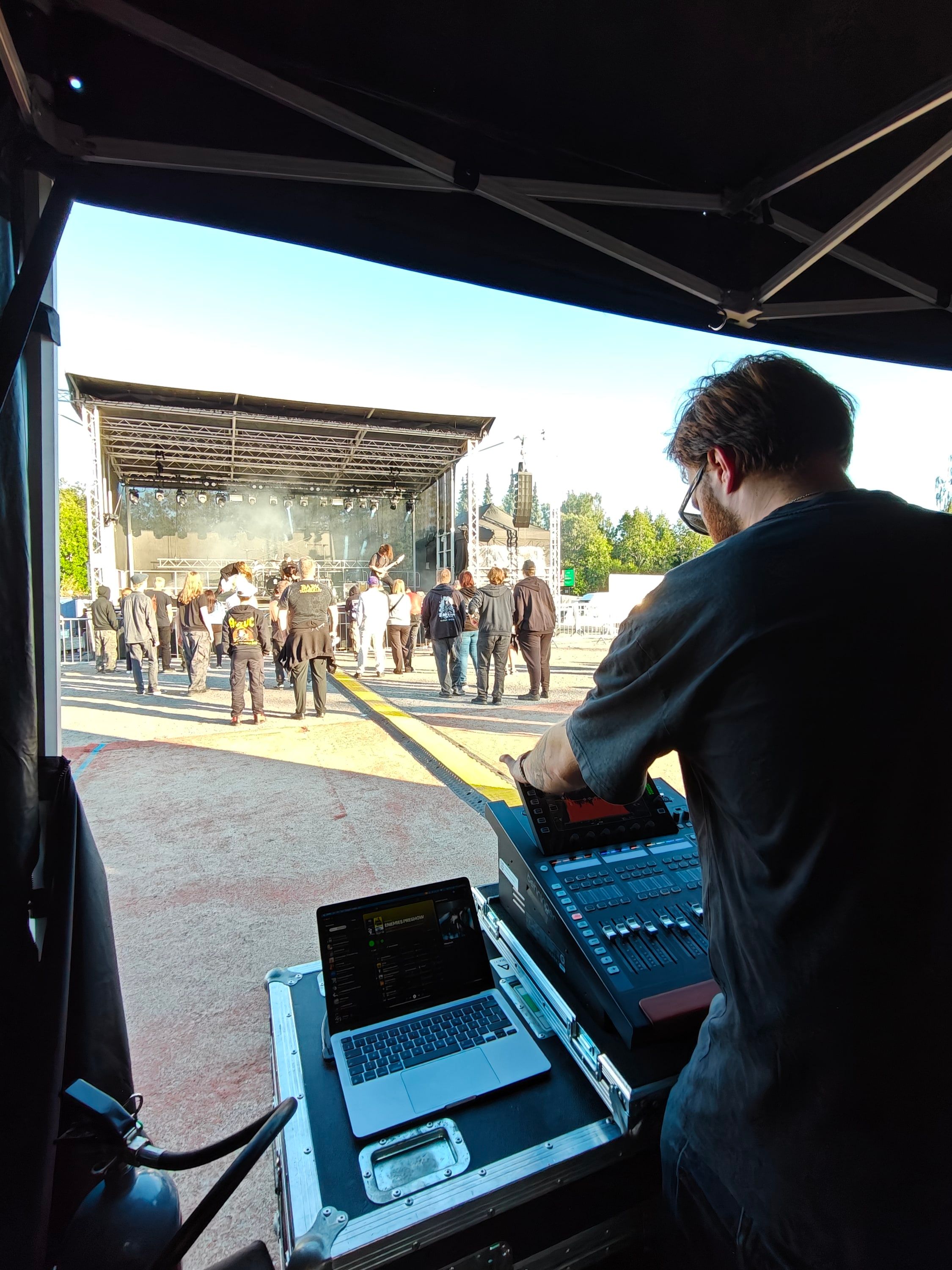 Akseli Kallunki mixing front of house from a tent at an outdoor festival, the crowd and stage ahead in evening sun.