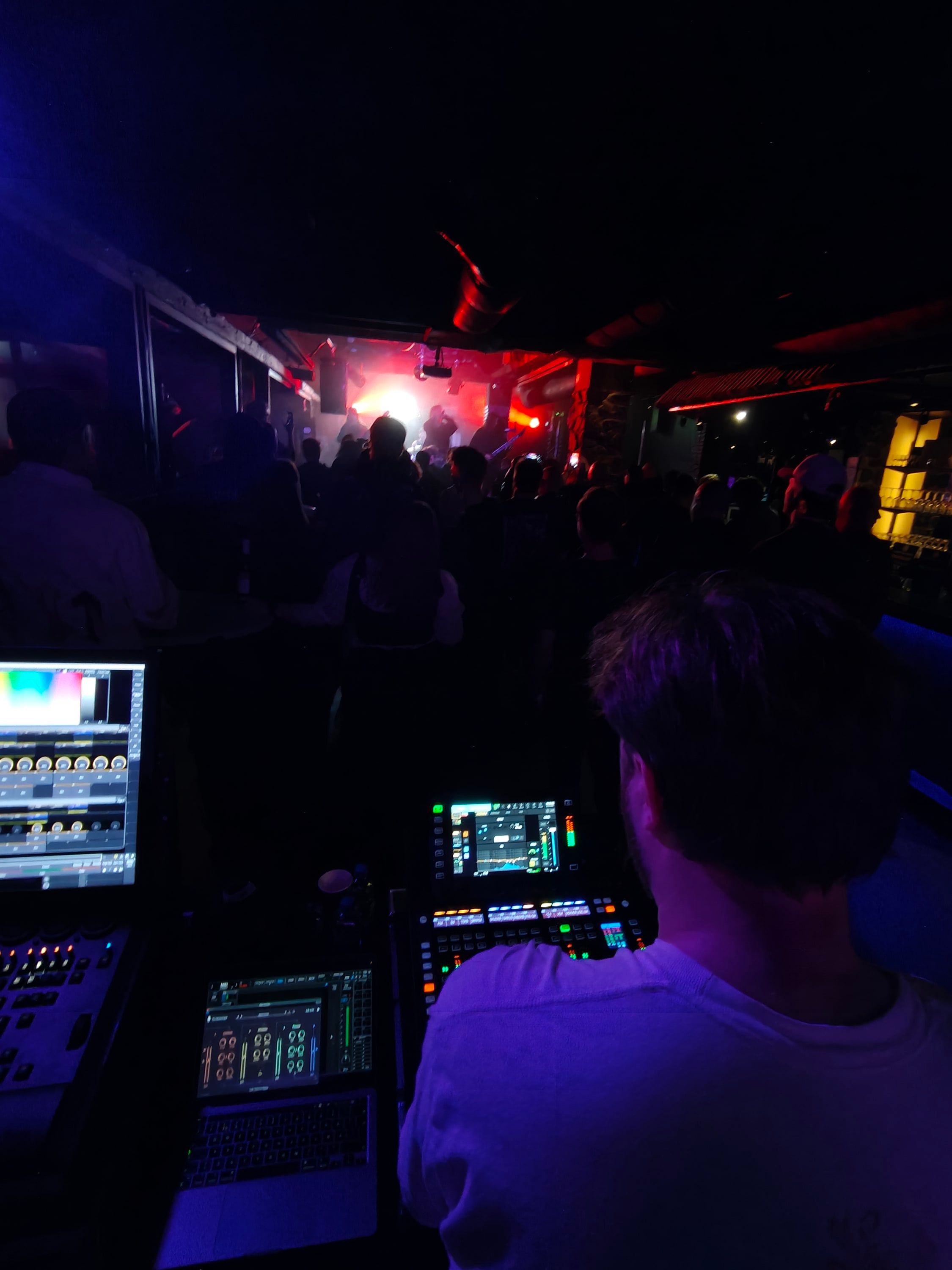 Over-the-shoulder view from the front-of-house desk: Akseli Kallunki at the console, the crowd and a red-lit stage ahead.